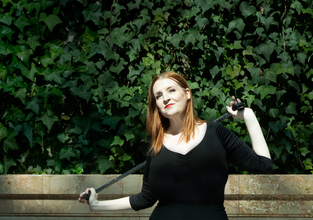 Portrait of Mary Fran, a disabled woman dressed in black standing in front of an ivy wall holding her cane.